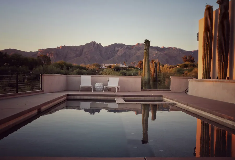 A still backyard pool reflecting tall saguaro cactus, with rugged desert mountains in the distance at dusk