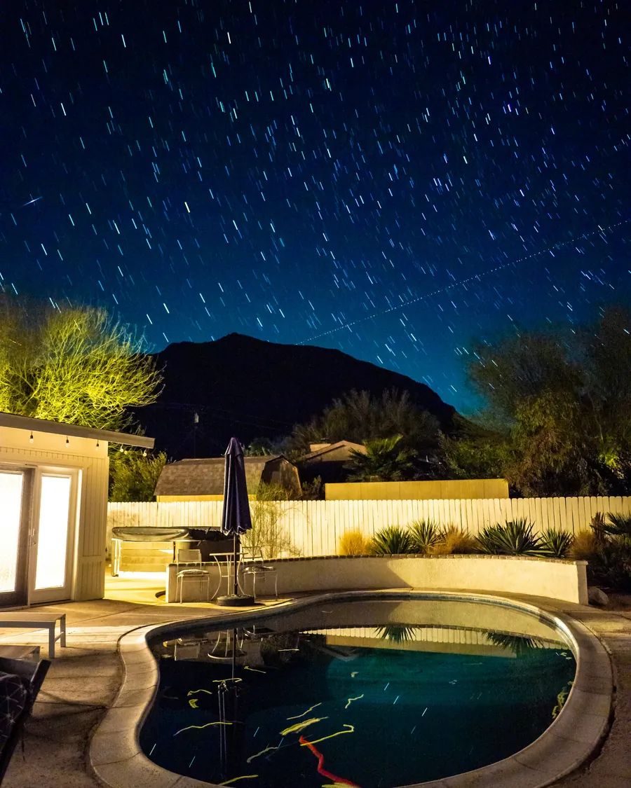 A lit backyard spa and patio at night under star trails, with a dark mountain behind